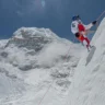 A climber in a red and white suit and blue helmet ascends a steep, snow-covered mountain slope, secured with orange climbing ropes. The rugged, icy peak and surrounding snow-covered ridges rise sharply against a clear blue sky, with a faint rainbow visible in the distance.