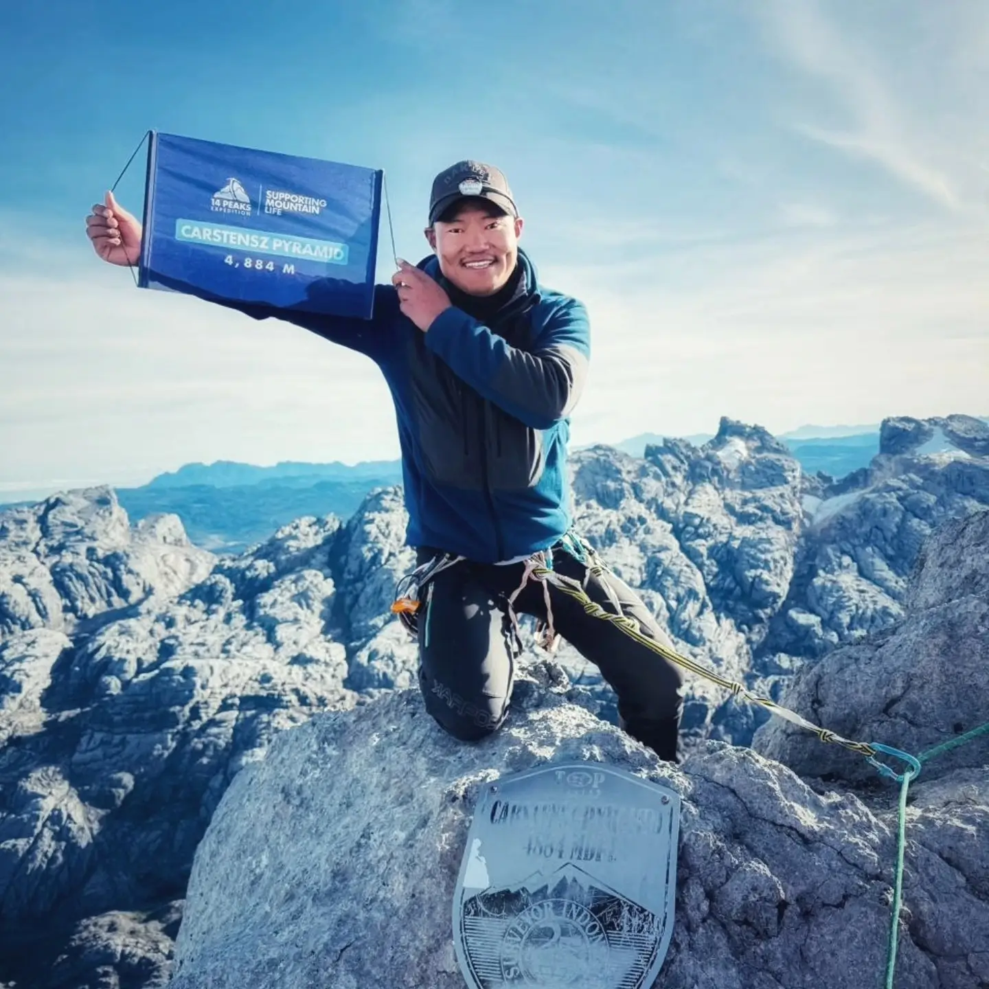A solo climber kneeling on the summit of Carstensz Pyramid, holding a sign that reads "CARSTENSZ PYRAMID 4,884 M" with the "14 Peaks" logo and "Supporting Life" text. The climber is equipped with climbing gear, and the background showcases a vast expanse of rocky mountains under a clear blue sky.
