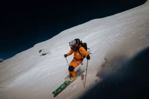 A skier in an orange suit and helmet is descending a steep, snow-covered slope at night, using ski poles for balance. The skier carries a backpack and leaves a trail in the snow, with a dark sky and shadowy mountains in the background.