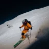 A skier in an orange suit and helmet is descending a steep, snow-covered slope at night, using ski poles for balance. The skier carries a backpack and leaves a trail in the snow, with a dark sky and shadowy mountains in the background.