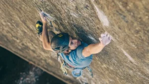 A rock climber in a helmet and harness ascends a steep, textured rock face, using both hands and feet to grip the surface, with climbing gear visible on their waist.
