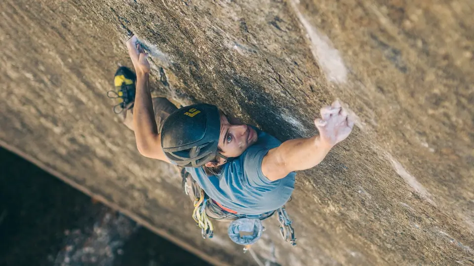 A rock climber in a helmet and harness ascends a steep, textured rock face, using both hands and feet to grip the surface, with climbing gear visible on their waist.