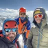 Three climbers wearing helmets and sunglasses pose for a selfie against a backdrop of snow-covered mountains under a partly cloudy sky. They are dressed in colorful mountaineering gear, including jackets and harnesses, with one holding an ice axe.