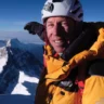 A person in a yellow and black mountaineering jacket and white helmet with "The North Face" logo, smiling and posing with gloved hands near snowy mountains under a clear blue sky