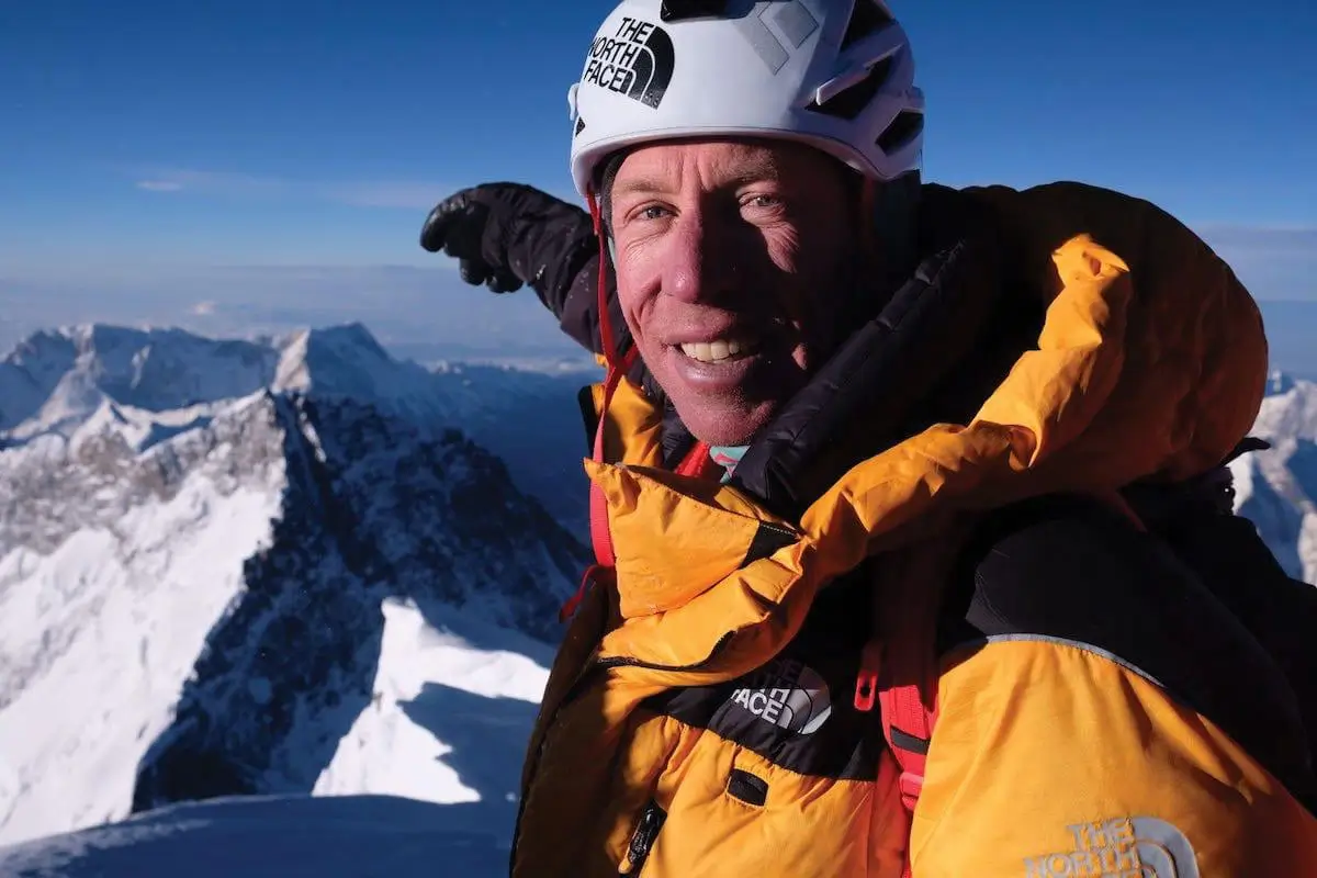 A person in a yellow and black mountaineering jacket and white helmet with "The North Face" logo, smiling and posing with gloved hands near snowy mountains under a clear blue sky
