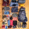 A neatly organized collection of camping and hiking gear laid out on a wooden floor. The items include a red rain jacket, a blue puffy jacket with a black face mask and sunglasses, a gray sleeping bag, a yellow tent, a purple sleeping pad, a large gray backpack, hiking poles, various clothing items (jackets, shirts, pants, underwear), a water bottle, a first aid kit, toiletries, a small pot, flip-flops, and trail running shoes.