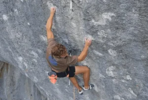 A person rock climbing on a steep, rugged cliff face, using both hands and feet to grip the rock. They are wearing a brown shirt, black shorts, and climbing shoes, with a harness and gear attached, including an orange chalk bag. A rope is secured to the harness and anchored to the rock above.