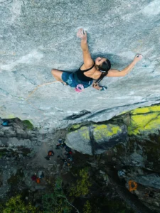 A rock climber ascends a steep granite cliff, gripping the rock face with both hands and feet. The climber is secured with an orange rope and wears a black top, blue shorts, and a chalk bag. Below, a group of people and camping gear are visible at the base of the cliff, surrounded by green vegetation and yellow lichen on the rocks. The scene is captured from an overhead perspective, highlighting the height and rugged terrain.