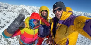 Three climbers in colorful mountaineering gear pose together on a snowy peak, with a stunning view of snow-covered mountains and a clear blue sky in the background.