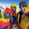 Three climbers in colorful mountaineering gear pose together on a snowy peak, with a stunning view of snow-covered mountains and a clear blue sky in the background.