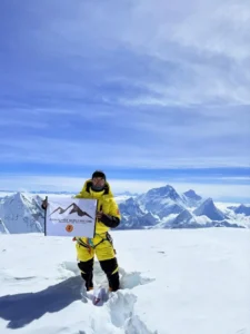 A person in a bright yellow mountaineering suit stands on a snowy mountain peak, holding a flag that reads "ANIMAL-FREE WORLD RECORD" with a mountain graphic and additional text. The background features a stunning view of snow-covered mountain ranges under a clear blue sky with some clouds.
