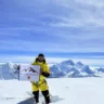 A person in a bright yellow mountaineering suit stands on a snowy mountain peak, holding a flag that reads "ANIMAL-FREE WORLD RECORD" with a mountain graphic and additional text. The background features a stunning view of snow-covered mountain ranges under a clear blue sky with some clouds.