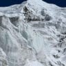 A close-up view of a rugged, snow-covered mountain landscape featuring steep ice formations and glaciers. The scene is dominated by white snow and ice against a deep blue sky, with rocky outcrops visible beneath the snow.