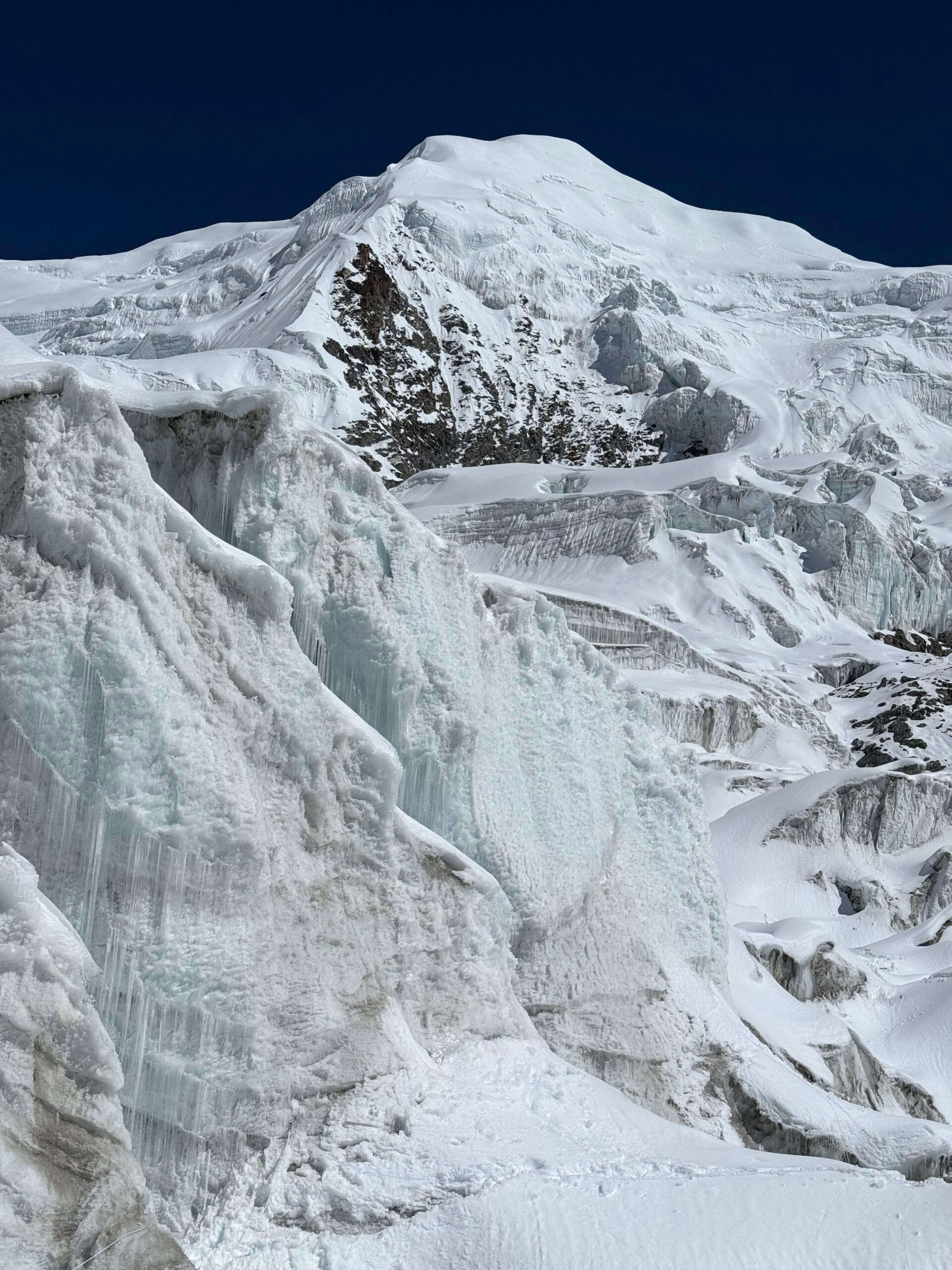 A close-up view of a rugged, snow-covered mountain landscape featuring steep ice formations and glaciers. The scene is dominated by white snow and ice against a deep blue sky, with rocky outcrops visible beneath the snow.