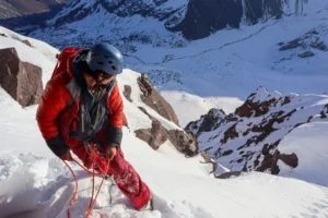 A climber in a red jacket and blue helmet ascends a steep, snow-covered mountain slope, secured with an orange rope. The background features rugged, snow-dusted peaks under a clear sky.