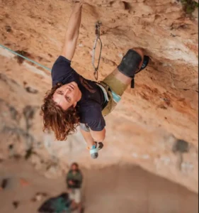 A rock climber in a dark shirt and green shorts hangs upside down from a steep rock face, secured by a rope, with another person visible below on the ground.