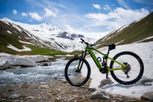 A green Pinarello electric mountain bike is parked on a rocky terrain near a flowing stream, with snow-covered mountains and green hills in the background under a bright blue sky with scattered clouds.