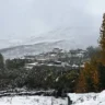 A snowy landscape featuring a cluster of traditional buildings with flat roofs, nestled on a hillside. The structures are partially covered in snow, surrounded by trees with autumn-colored leaves. Snow-covered mountains rise in the background under a cloudy, overcast sky.