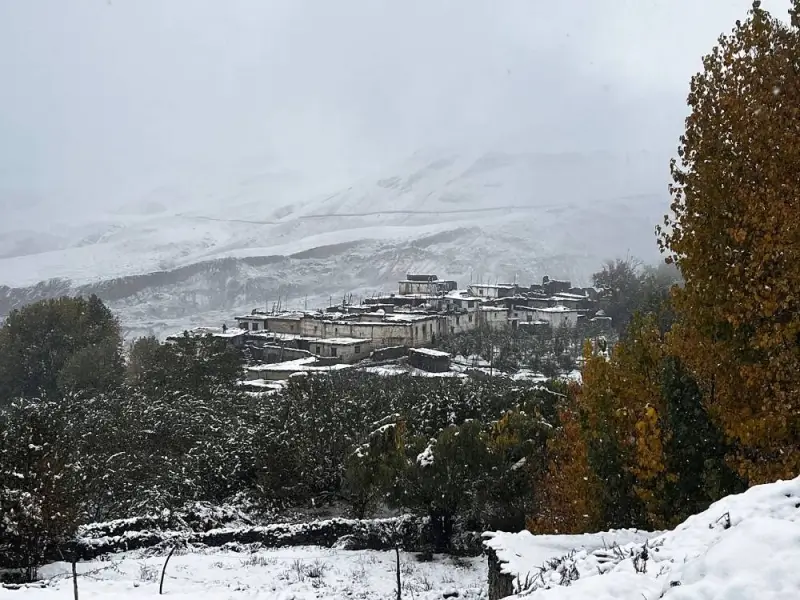 A snowy landscape featuring a cluster of traditional buildings with flat roofs, nestled on a hillside. The structures are partially covered in snow, surrounded by trees with autumn-colored leaves. Snow-covered mountains rise in the background under a cloudy, overcast sky.