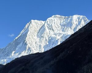 A majestic snow-covered mountain peak rises sharply against a clear blue sky, with rugged ridges and icy slopes illuminated by bright sunlight. A dark, rocky foreground ridge contrasts with the pristine white snow.