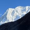 A majestic snow-covered mountain peak rises sharply against a clear blue sky, with rugged ridges and icy slopes illuminated by bright sunlight. A dark, rocky foreground ridge contrasts with the pristine white snow.