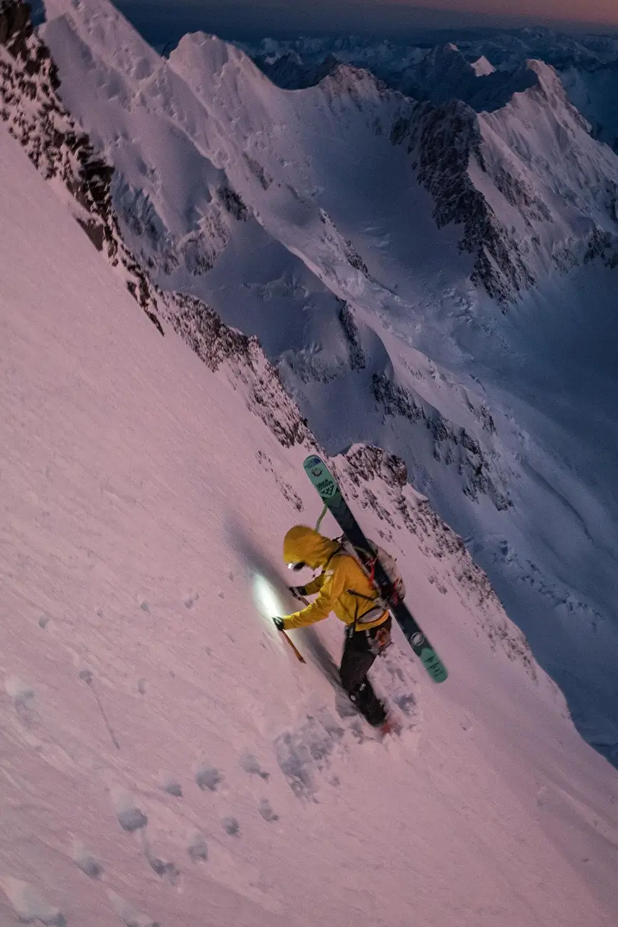 A skier in a yellow jacket climbs a steep, snow-covered mountain slope at dawn or dusk, using a headlamp to illuminate the path ahead. The skier carries skis on their back and holds an ice axe in one hand. The vast, rugged mountain landscape stretches into the distance under a gradient sky transitioning from pink to blue.