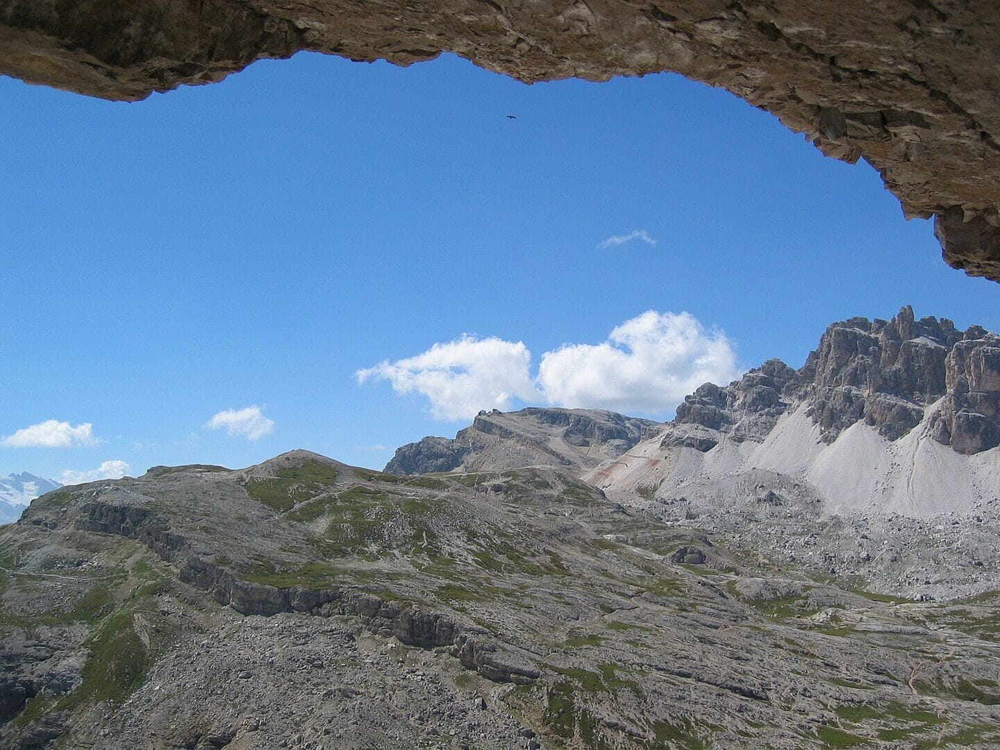 View from inside a rocky cave opening, framing a vast alpine landscape with jagged mountain peaks, patches of green vegetation, and scattered rocks under a bright blue sky with fluffy white clouds. A small bird is visible flying in the distance.