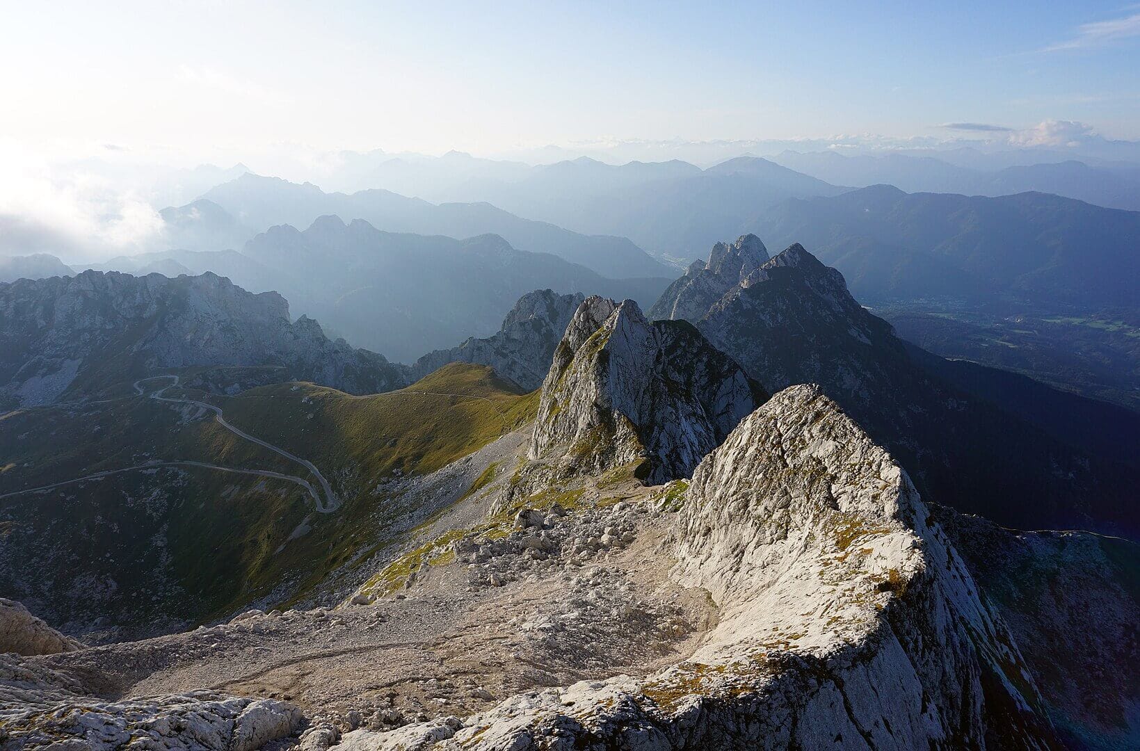 Panoramic sunrise or sunset view from a high summit in the Julian Alps, Slovenia. In the foreground, jagged white limestone ridges and a winding mountain road drop steeply into shadowed valleys. Layer upon layer of distant mountain ranges fade into blue haze under a glowing sky.