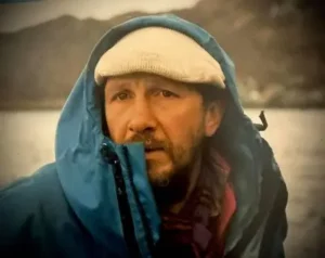 A bearded man wearing a light-colored knit cap and a blue hooded jacket pulled up around his face, standing outdoors near a body of water with a blurred natural landscape in the background.