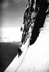 A dramatic black-and-white photograph of two climbers on the near-vertical northeast face of the Eiger. One climber is high above, dangling precariously from a rope against the sheer rock wall, while the second climber, far below, stands on a steep snow slope holding the rope, looking up at their partner. The vast drop and clouds below emphasize the extreme exposure of the Eiger Nordwand.