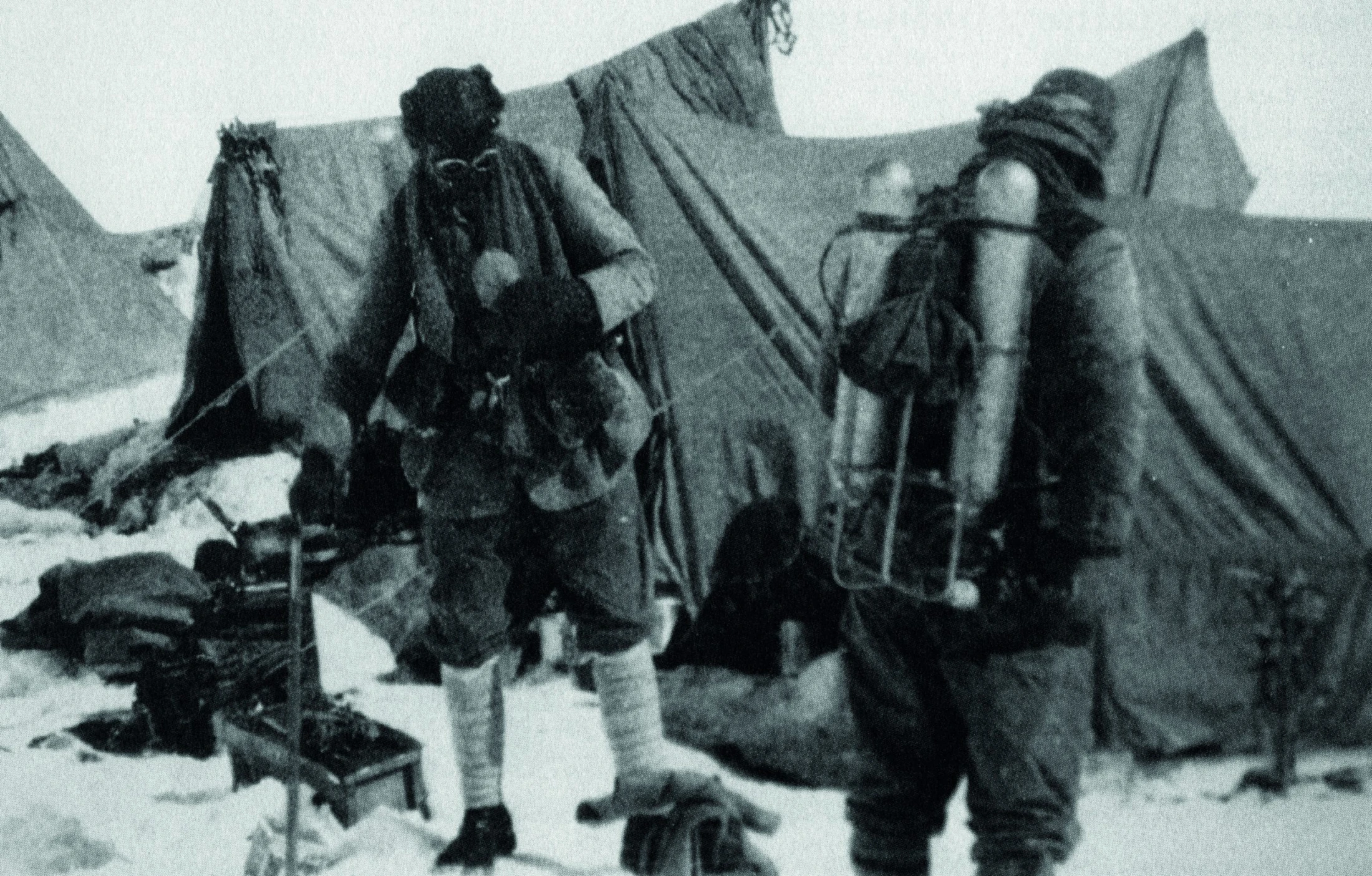 Black-and-white photograph from the 1924 British Mount Everest Expedition showing George Mallory (left) and Andrew “Sandy” Irvine (right) at or near the North Col. Both are dressed in heavy layered wool clothing, hobnail boots, and early climbing gear, carrying ice axes and wearing wide-brimmed hats. They are roped together on snowy, rocky terrain with tent fabric visible behind them. This is the last known photograph of the pair before their fatal summit attempt.