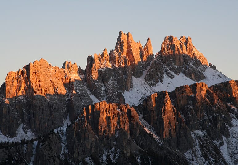 Jagged, snow-dusted mountain peaks glow with warm golden light from the setting or rising sun, casting dramatic shadows across the rugged rock faces under a clear pale blue sky.