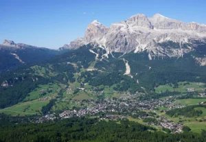 Aerial view of a picturesque alpine village nestled in a lush green valley, surrounded by dense forests and rolling meadows, with majestic snow-capped mountains rising dramatically in the background under a clear blue sky.
