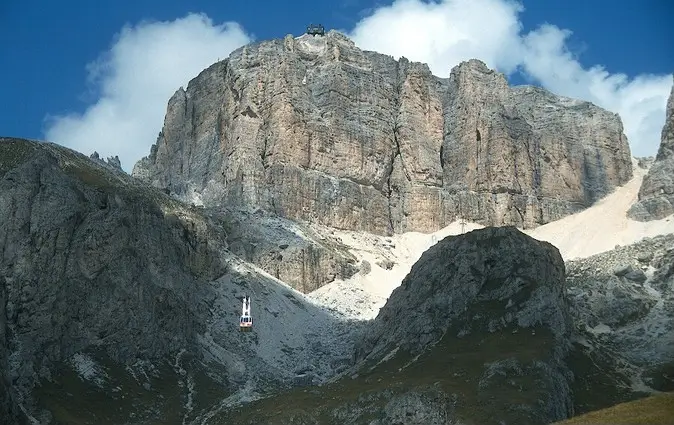 A red cable car ascends toward a small station perched atop a sheer, rugged mountain peak, surrounded by dramatic rocky cliffs and patches of snow under a partly cloudy blue sky.