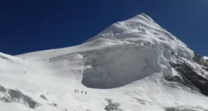 A group of climbers ascends a steep, snow-covered slope toward a towering, jagged mountain peak under a clear blue sky. The mountain features prominent ice cliffs and crevasses, with the climbers appearing small against the vast, icy landscape.