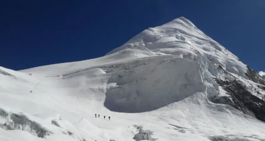 A group of climbers ascends a steep, snow-covered slope toward a towering, jagged mountain peak under a clear blue sky. The mountain features prominent ice cliffs and crevasses, with the climbers appearing small against the vast, icy landscape.