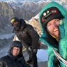 Three climbers in heavy winter gear pose for a selfie on a rocky, snow-covered ridge with a dramatic backdrop of jagged, snow-capped mountain peaks under a clear blue sky.