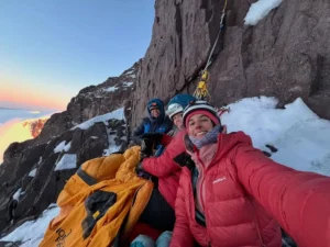 Three smiling female climbers in heavy winter gear and helmets take a sunrise selfie while bivouacked on a portaledge hanging from a steep granite wall high on Cerro Steffen, Patagonia. An orange sleeping bag and fixed ropes are visible against a backdrop of snow-covered rock and glowing dawn light.