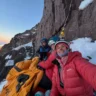 Three smiling female climbers in heavy winter gear and helmets take a sunrise selfie while bivouacked on a portaledge hanging from a steep granite wall high on Cerro Steffen, Patagonia. An orange sleeping bag and fixed ropes are visible against a backdrop of snow-covered rock and glowing dawn light.