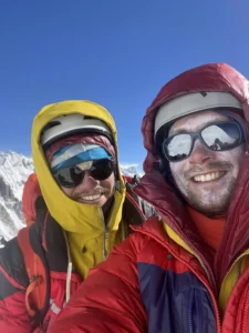Two smiling climbers in heavy mountaineering gear (one in a bright yellow hooded jacket, the other in red and purple) take a selfie high on a snowy mountain ridge under a deep blue sky, both wearing helmets, glacier glasses, and frost-covered faces, with distant peaks visible behind them.