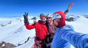 Three smiling climbers in helmets and mountaineering gear pose for a selfie on a snowy mountain summit. One makes a peace sign, another waves, and the third takes the photo. The background features a stunning expanse of snow-covered peaks under a clear blue sky.