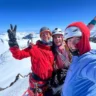 Three smiling climbers in helmets and mountaineering gear pose for a selfie on a snowy mountain summit. One makes a peace sign, another waves, and the third takes the photo. The background features a stunning expanse of snow-covered peaks under a clear blue sky.