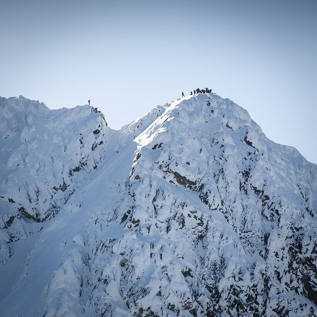 A line of mountaineers in colorful winter gear ascends the final snow-covered ridge toward a sharp, sunlit summit, silhouetted against a pale winter sky. The steep, pristine slopes drop away dramatically on both sides, emphasizing the exposed and exhilarating high-alpine environment.