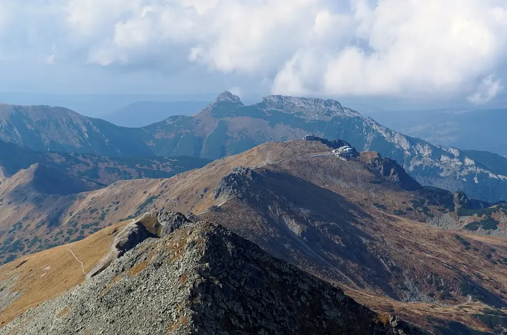 Panoramic view from a high alpine ridge, looking across barren, rocky slopes toward distant snow-dusted peaks. A small cluster of buildings, likely a mountain observatory or cable car station, sits perched on a nearby summit under a partly cloudy sky.