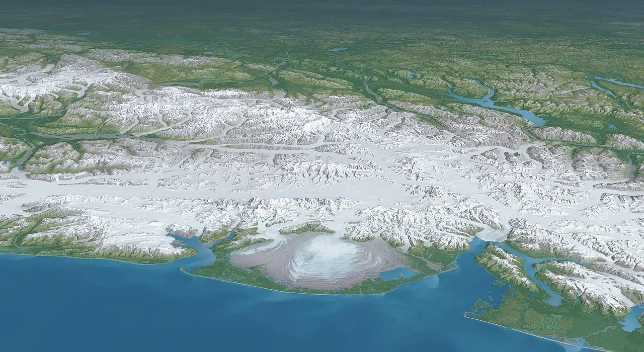 An expansive aerial or satellite view of the Malaspina Glacier in southeastern Alaska, the world's largest piedmont glacier. The massive central lobe of swirling, folded ice spreads out onto a coastal plain, surrounded by intricate looped moraines carrying debris. Snow-covered mountain peaks rise in the background, with green forested areas and lakes visible on the periphery, bordering the deep blue Gulf of Alaska in the foreground.