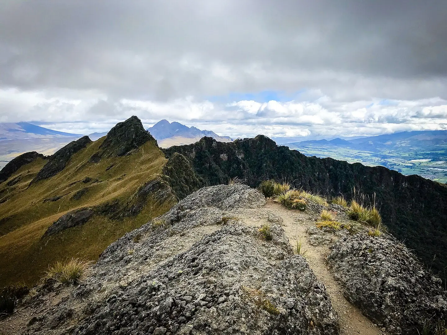 Dramatic high-altitude view from a rocky mountain ridge with a narrow dirt trail running along the crest. Steep, grassy slopes drop away on the left, while dark forested cliffs fall sharply on the right. In the distance, layered mountain peaks rise beneath a moody, cloud-filled sky, with low-lying clouds partially obscuring the valley far below.1.7sFast