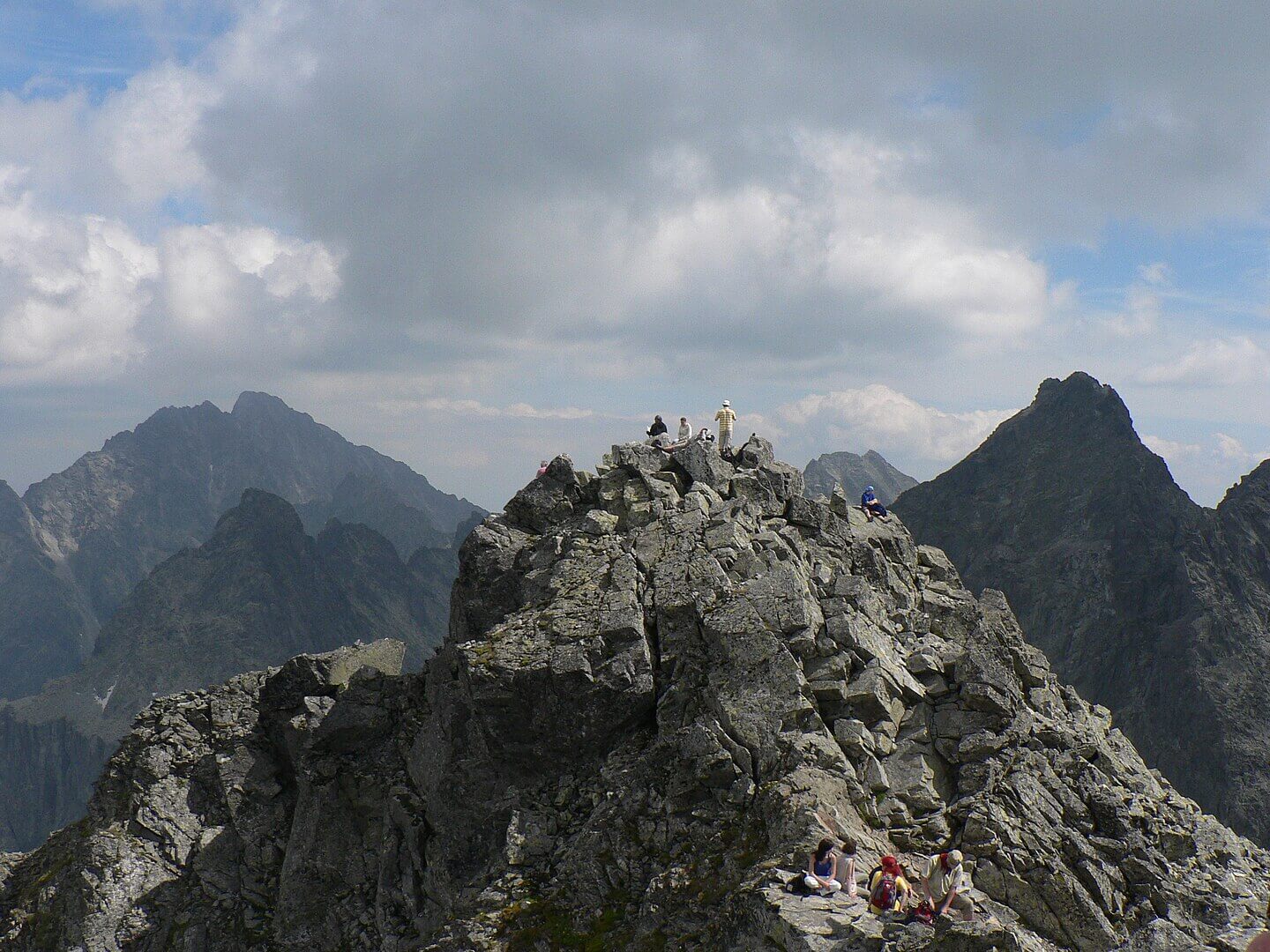 A group of hikers perched on the jagged, rocky summit of a high mountain peak, resting and taking in the breathtaking view. Dramatic alpine ridges and distant pointed summits stretch across the horizon beneath a sky filled with towering clouds.