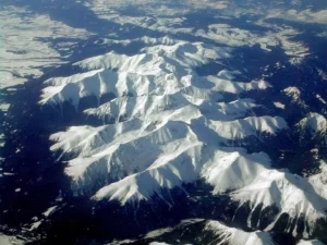 Aerial view of rugged, snow-capped mountain peaks bathed in sunlight, with sharp ridges and deep valleys casting dramatic shadows. Dark evergreen forests blanket the lower slopes, contrasting against the bright white snow cover under a clear blue sky.