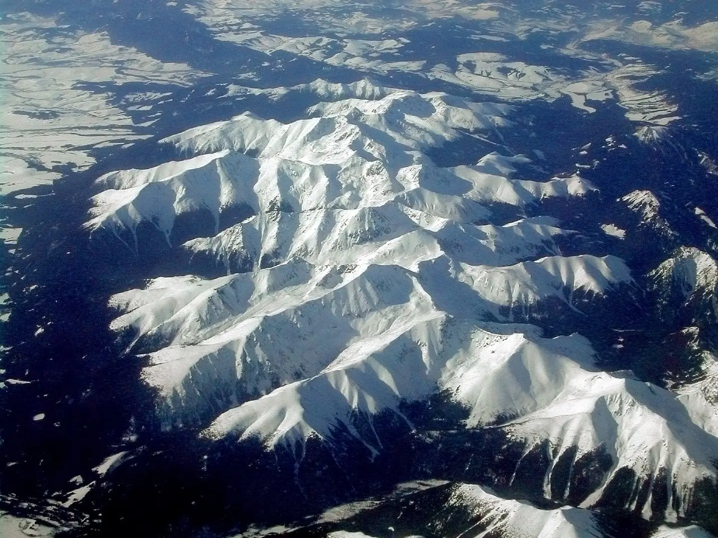Aerial view of rugged, snow-capped mountain peaks bathed in sunlight, with sharp ridges and deep valleys casting dramatic shadows. Dark evergreen forests blanket the lower slopes, contrasting against the bright white snow cover under a clear blue sky.