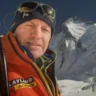 A middle-aged mountaineer takes a selfie in high-altitude conditions, squinting and smiling slightly against bright sunlight, wearing a dark beanie, glacier sunglasses pushed up on his forehead, and a bright orange down jacket with "KAYLAND" and "ACERBIS" sponsor logos on the chest. An ice axe is strapped to his backpack over his left shoulder, and the iconic snow-covered peak of Mount Everest looms prominently in the background under a clear blue sky.